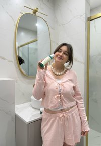 Bottiglia di shampoo verde in un bagno, tenuta da una persona che indossa un completo a due pezzi rosa chiaro a costine con dettagli a coulisse e perle.
