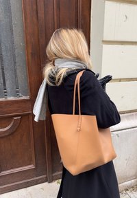 Sac fourre-tout en cuir fauve au design minimaliste, présentant une texture lisse et une seule lanière. Le sac comporte des coutures inclinées pour la structure.