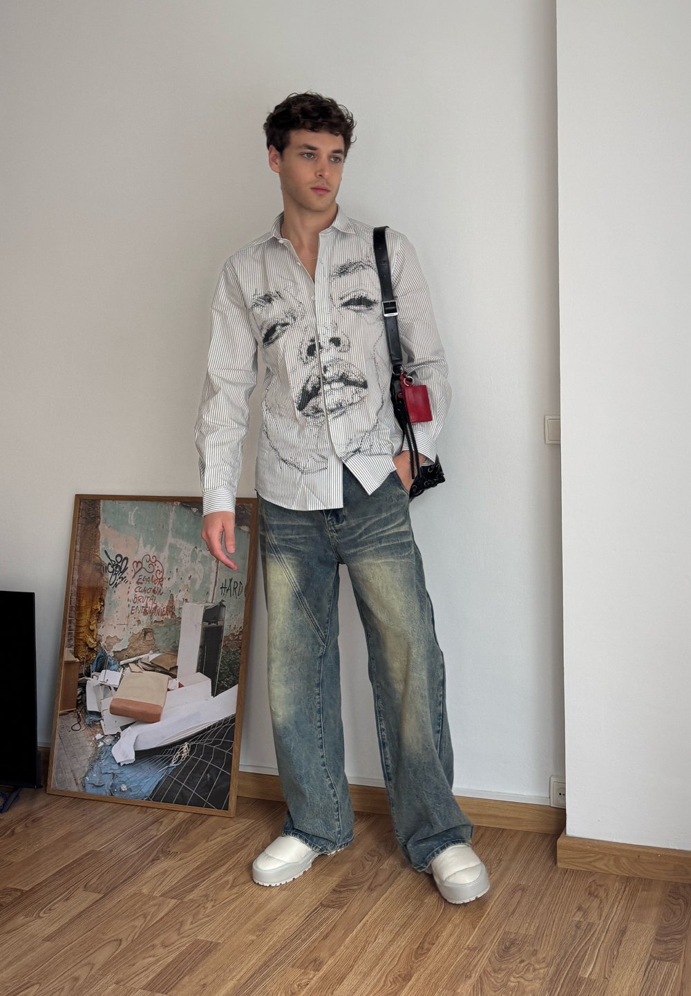 Camicia bianca a righe con un volto stampato, abbinata a jeans baggy blu chiaro e scarpe bianche chunky. Il modello tiene in mano una borsa nera e rossa.