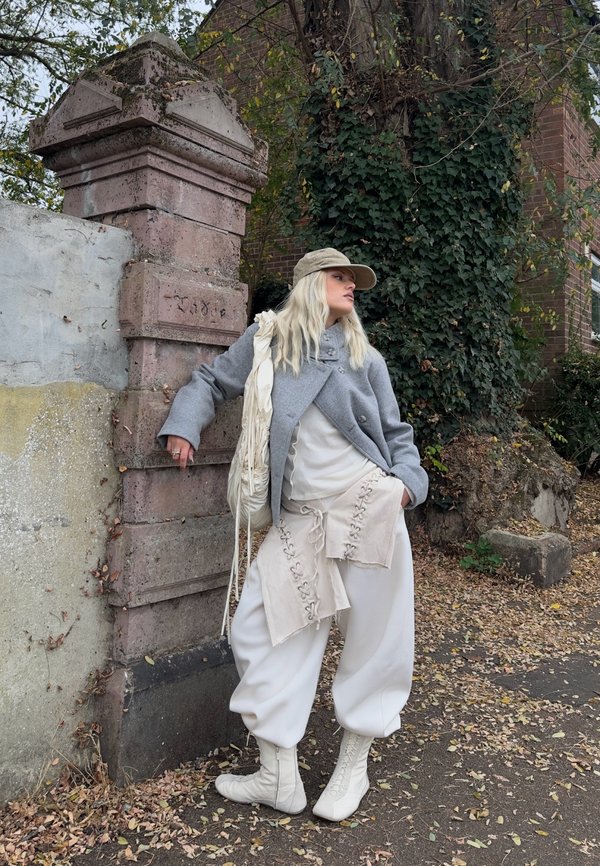 Una mujer rubia con gorra, abrigo gris, capas crema y botas blancas posa junto a una pared de ladrillos cubierta de hiedra al aire libre.
