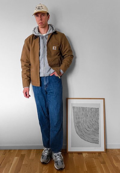 Chaqueta marrón con cremallera sobre una sudadera gris con capucha, vaqueros de mezclilla azules y zapatillas grises. Una obra de arte en blanco y negro enmarcada está en el suelo al lado de él.