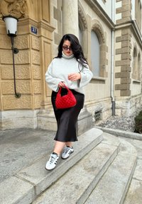 Υπερμεγέθη γκρι ριγέ πουλόβερ, μαύρη σατέν φούστα, κόκκινη πλεκτή τσάντα και λευκά αθλητικά παπούτσια με μαύρες λεπτομέρειες; βήματα και αρχιτεκτονική από πέτρα στο φόντο.