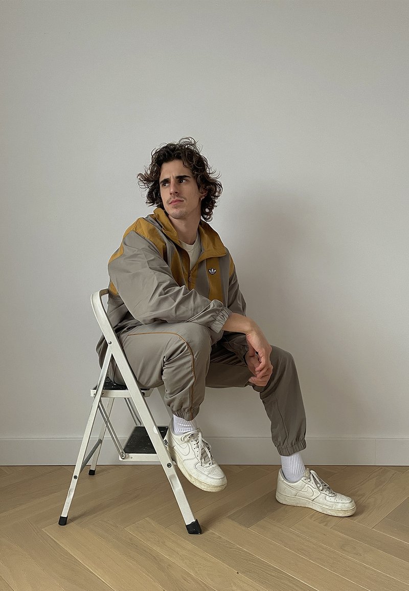 Šviesiai pilkas treningas su garstyčių geltonumo priekiu, su elastinėmis kelnėmis ir baltomis sportinėmis bateliais. Puošnios garbanos, sėdi ant sulankstomo stalo.