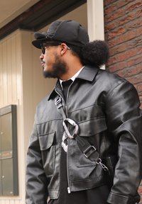 Veste en cuir noir avec deux poches avant, une fermeture éclair, et une sangle amovible avec un détail de mousqueton argenté. Portée sur une chemise foncée.