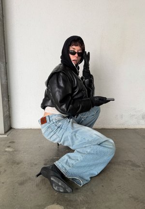 Chaqueta bomber negra de cuero recortada, sudadera con capucha gris, gafas de sol oscuras, guantes negros, jeans azules holgados y tacones de aguja negros. Fondo minimalista.