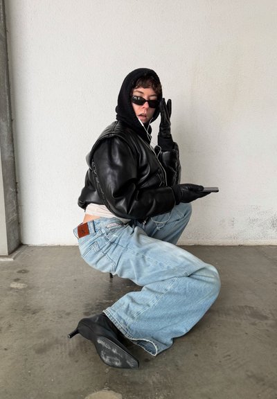 Chaqueta bomber negra de cuero recortada, sudadera con capucha gris, gafas de sol oscuras, guantes negros, jeans azules holgados y tacones de aguja negros. Fondo minimalista.