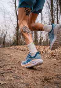 Ténis de corrida azul claro com detalhes laranja, solas texturizadas e um logótipo de marca reconhecível, usados com meias brancas até ao tornozelo.