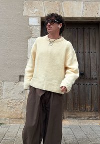 Bărbat purtând un pulover crem supradimensionat, pantaloni maro cu dungi fine, colier în formă de inimă, ochelari de soare și căști wireless, stând lângă o ușă din lemn.