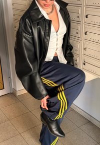 Άτομο που φοράει μαύρο δερμάτινο μπουφάν, λευκό πουκάμισο με κουμπιά, μπλε σκούρο αθλητικό παντελόνι με κίτρινες ρίγες και μαύρα μποτάκια, ρυθμίζει το μπατζάκι του παντελονιού κοντά σε θυρίδες ταχυδρομείου.