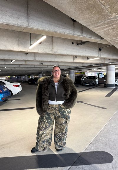 Chaqueta verde oscuro corta con mangas peludas, combinada con pantalones cargo de camuflaje. Ambientado en una estructura de estacionamiento de concreto con varios coches.