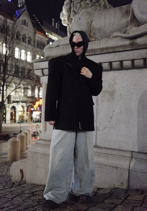 Jachetă oversized de culoare neagră cu glugă și accesorii din metal argintiu, purtată cu pantaloni evazați gri. Decorul este urban, pe timp de noapte, cu arhitectură din piatră.