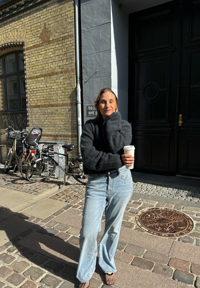 Suéter negro de gran tamaño, jeans de pierna ancha de color azul claro, sosteniendo una taza de café; el fondo presenta una pared de ladrillo y bicicletas.