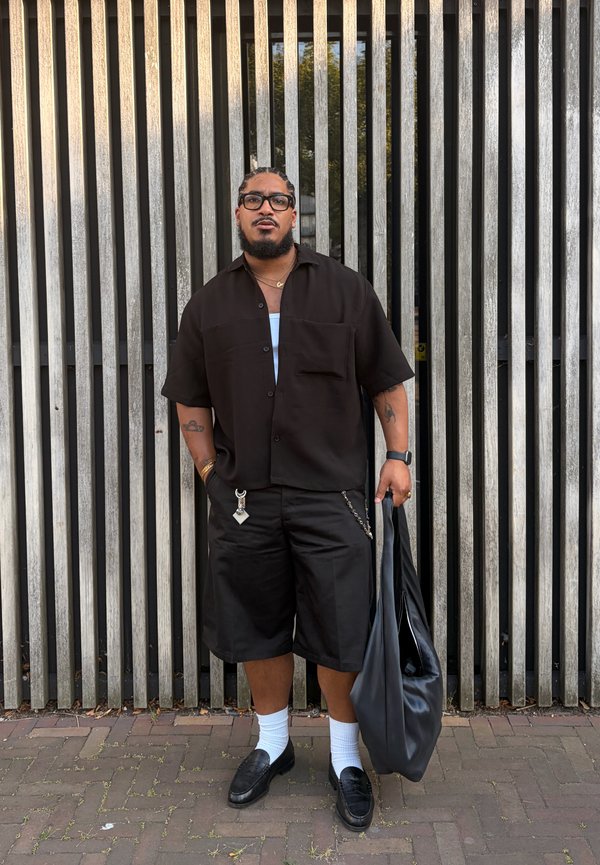 Man with glasses wearing black shirt and shorts, white socks, black loafers, holding large black bag, standing by vertical metal bars.