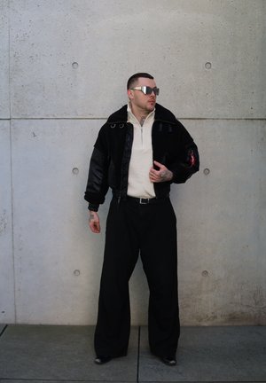 Chaqueta de shearling negro y cuero, suéter cremoso con cremallera, pantalones negros holgados, gafas de sol reflectantes de gran tamaño, tatuajes visibles en los brazos.