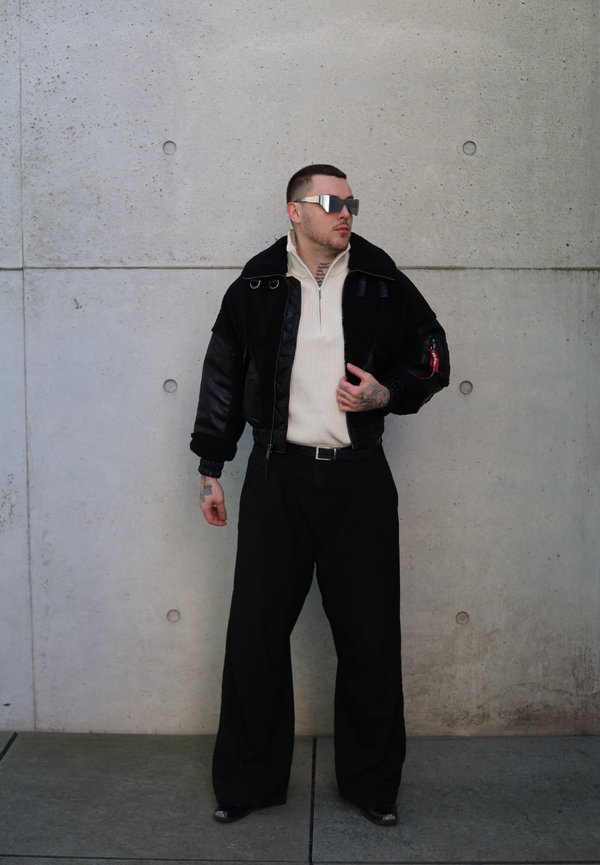 Casaco preto de shearling e pele, suéter creme com fecho éclair, calças soltas pretas, óculos de sol reflexivos oversized, tatuagens visíveis nos braços.