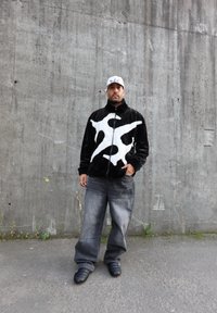 Juodas pūkuotas švarkas su baltu abstrakčiu piešiniu, derinamas su laisvais pilkais džinsais ir juodais slip-on batais, prieš betono sieną.