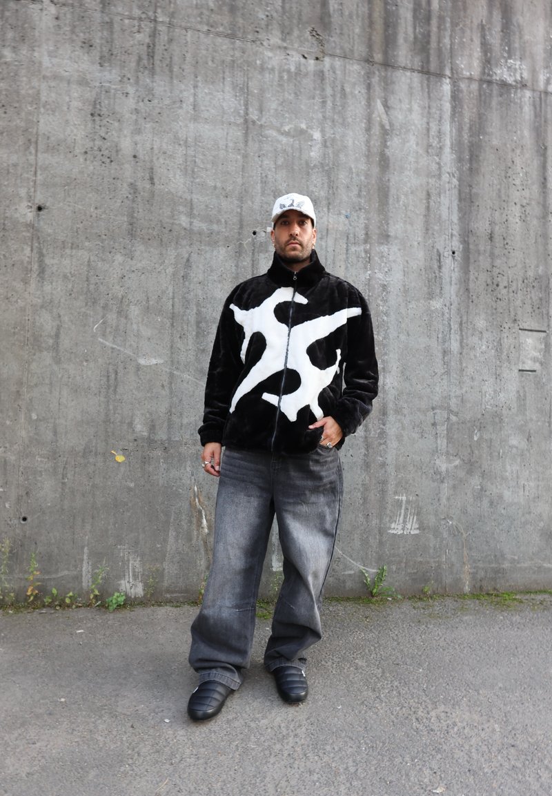 Veste en peluche noire avec un motif abstrait blanc, associée à un jean gris ample et des chaussures noires à enfiler, le tout devant un mur en béton.