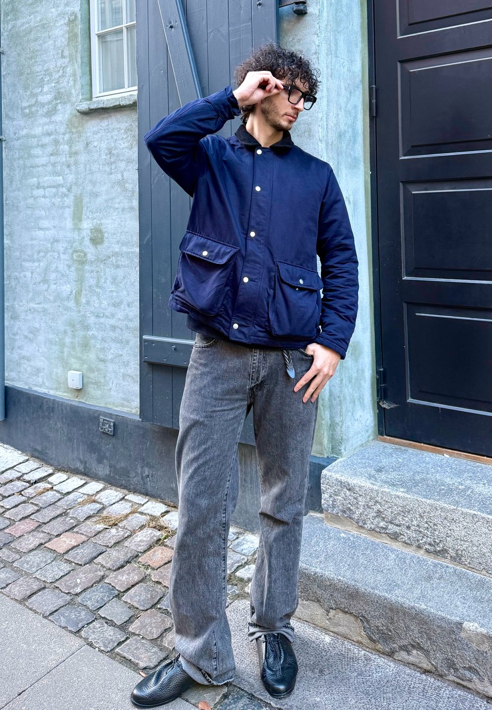 Veste matelassée marine avec deux poches avant et boutons-pression, portée avec un jean gris à jambes larges et des chaussures en cuir noir sur une rue pavée.