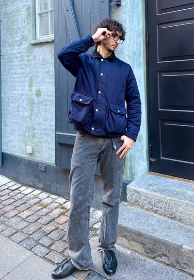 Chaqueta acolchada azul marino con dos bolsillos delanteros y botones a presión, combinada con jeans de pierna ancha grises y zapatos de cuero negros sobre una calle empedrada.