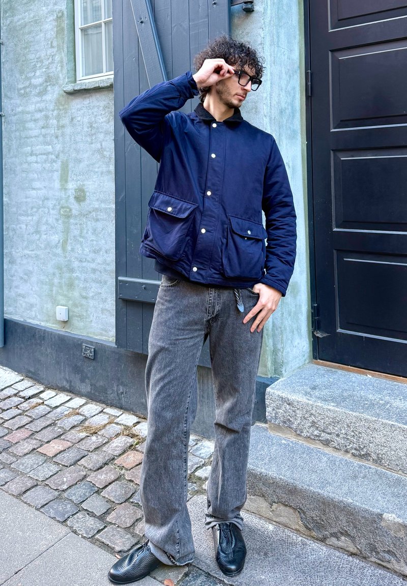 Veste matelassée marine avec deux poches avant et boutons-pression, portée avec un jean gris à jambes larges et des chaussures en cuir noir sur une rue pavée.