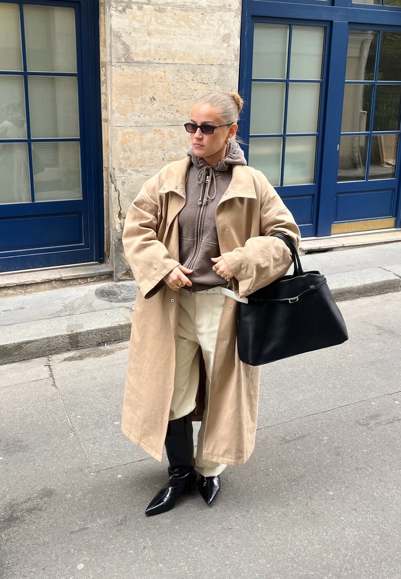 Femme portant un trench-coat beige, un sweat à capuche marron, un pantalon crème, des bottes noires et des lunettes de soleil, portant un grand sac à main noir dans une rue de la ville.