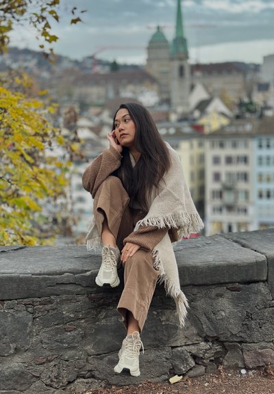 Suéter beige tejido, pantalones de pana marrones, chal claro con flecos y zapatillas beige. El fondo muestra un paisaje urbano con torres de iglesias.