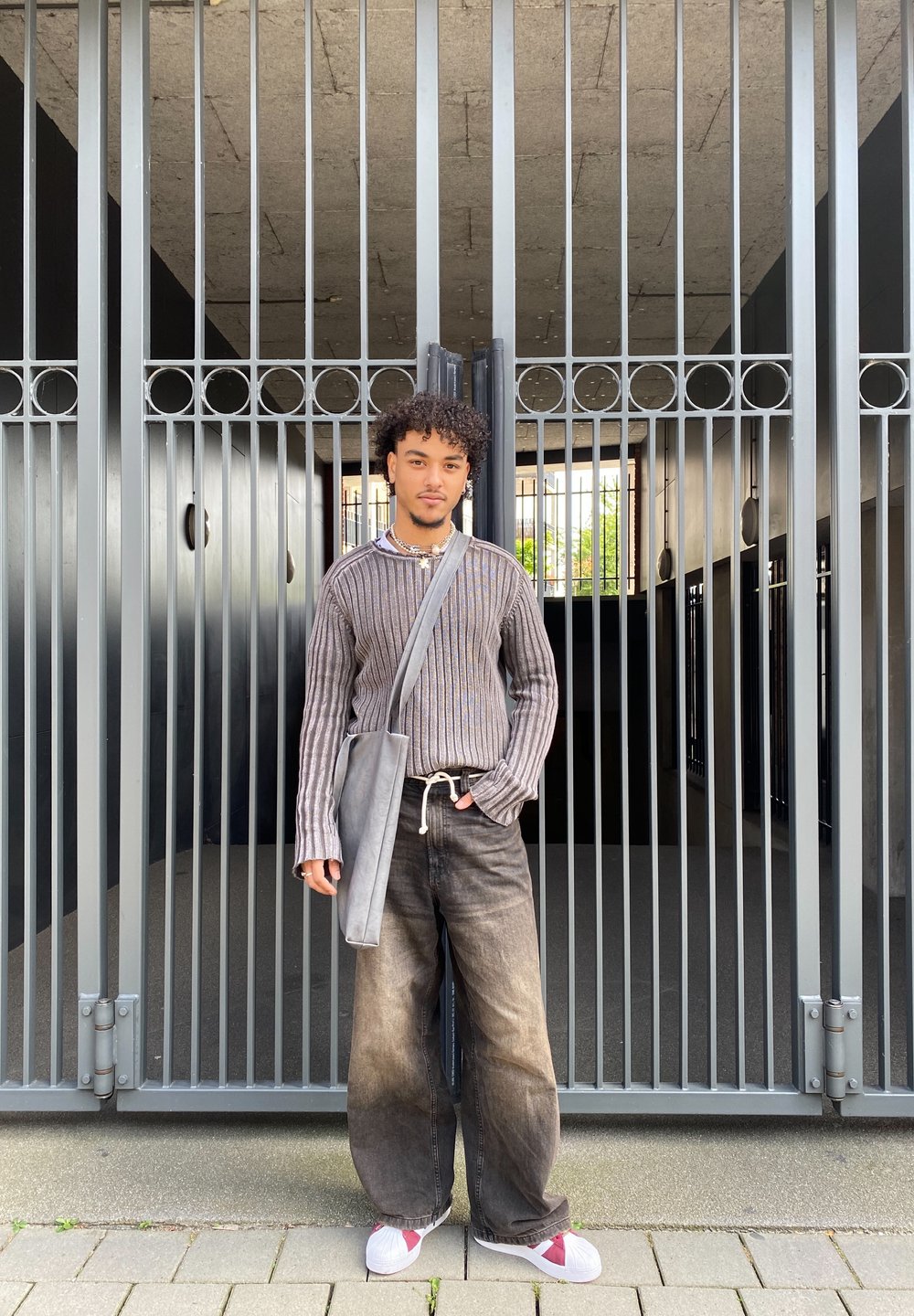 Grey vertically striped sweater, loose dark jeans, white shoes with red accents, grey shoulder bag, standing against a metal gate.