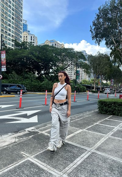 Conjunto informal que incluye una blusa sin mangas blanca y pantalones holgados gris claro. Zapatillas gris oscuro. Entorno urbano con vegetación y edificios de fondo.