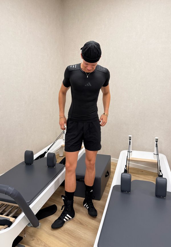 Schwarzes, enganliegendes Sportshirt mit strukturierten Schulterakzenten, kombiniert mit schwarzen Shorts und Schuhwerk mit weißen Streifen. Ausrüstung im Hintergrund.