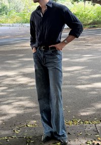 Granatowa koszula z długim rękawem, luźne niebieskie jeansy, czarny pasek i ciemne buty; stoi na ulicy z roślinnością w tle.