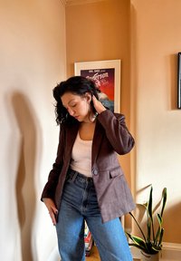 Smeđi antilop sako s jednim dugmetom, krojen s naglaskom na fit, i dva bočna džepa, kombiniran s trapericama visokog struka u plavoj boji. U pozadini se nalazi biljka.