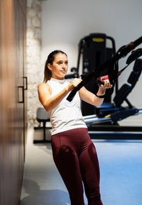 Insegnante di fitness che utilizza cinghie di sospensione nere, indossando una maglietta senza maniche bianca e leggings bordeaux, in una palestra moderna con pareti in legno e pietra.