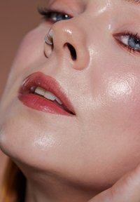 Nærbilde av et ansikt med glatt, glødende hud, en sølvfarget nesering, myke rosa lepper og subtil rouge. Blå øyne rammet inn av lange vipper.