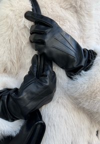 Guantes de cuero negro con detalles de costuras verticales, sosteniendo una correa de cuero negro, sobre un fondo de piel blanca suave.