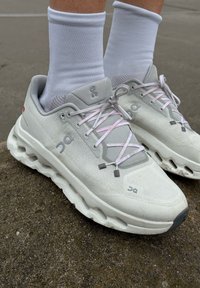 Scarpe da ginnastica grigio chiaro con lacci rosa e tomaia in mesh strutturato. Presentano una suola bianca imbottita con segmenti ritagliati per l'assorbimento degli urti.