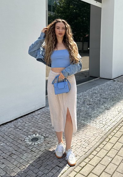 Top corto azul, falda beige clara con abertura lateral, chaqueta de mezclilla, bolso azul claro y zapatillas blancas con suelas de goma en un camino de adoquines.