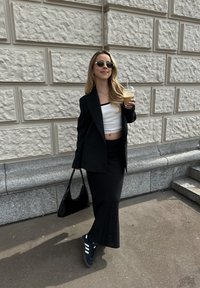 Crni sako, bijeli cropped top, crna maxi suknja, crne tenisice. Drži ledeni napitak. Glatka pozadina zida, opušteno urbano okruženje.