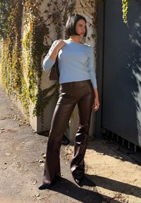 Svijetloplava dukserica, smešane kožne flare hlače, crne cipele s visokim potpeticama, stojeći uz teksturirani zid s zelenilom.