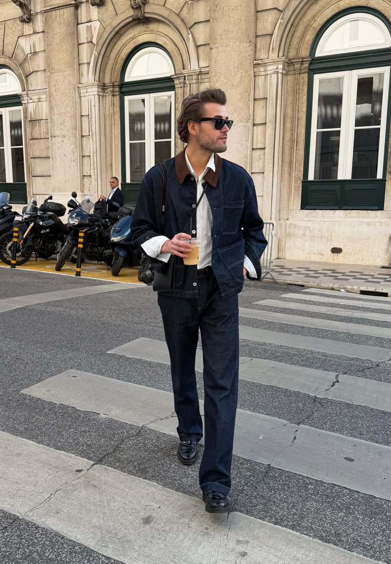 Mann mit Sonnenbrille und dunklem Denim-Outfit, der eine Kaffeetasse hält und eine Straße mit Motorrädern und historischem Gebäude im Hintergrund überquert.