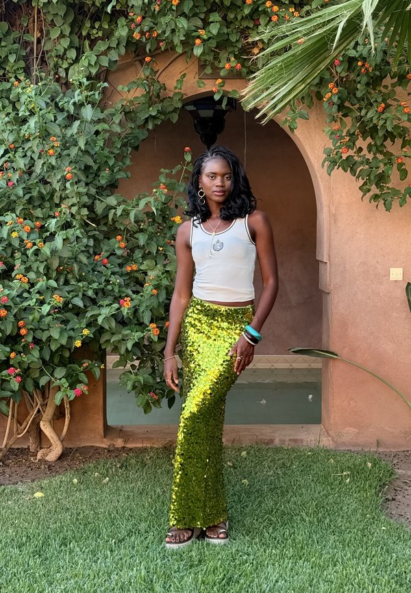 Groene paillettenrok met een aansluitende silhouet, gecombineerd met een witte tanktop. Accessoires zijn onder andere armbanden en een hanger ketting.