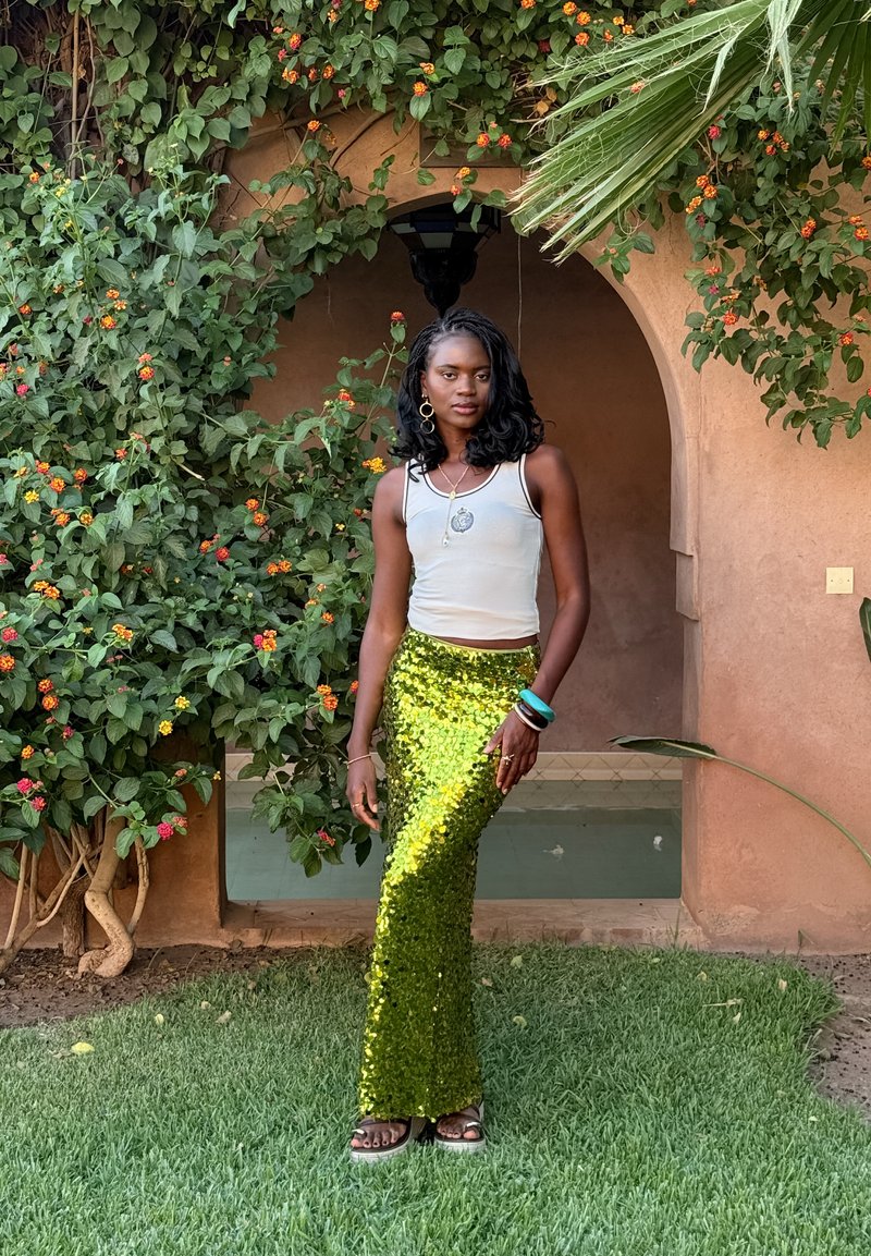 Jupe verte à sequins avec une silhouette ajustée, associée à un débardeur blanc. Les accessoires comprennent des bracelets et un collier avec pendentif.