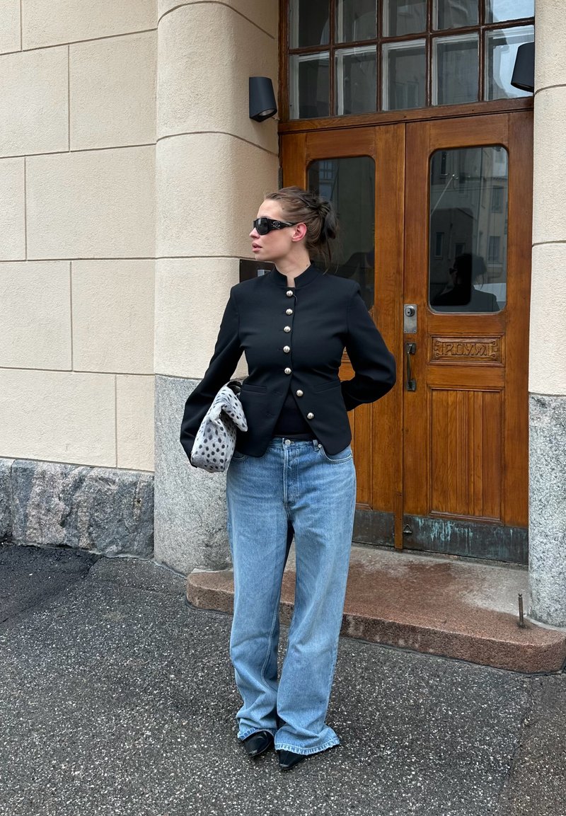 Mujer con chaqueta negra abotonada y gafas de sol sosteniendo una bolsa con estampado de manchas, de pie junto a una puerta de madera sobre suelo de piedra y asfalto.