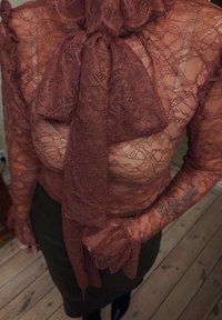 Kanten blouse in diepe roestkleur met een hoge kraag en strikdetail. Doorzichtige, bloemrijke kanten textuur met lange mouwen en gerimpelde manchetten.