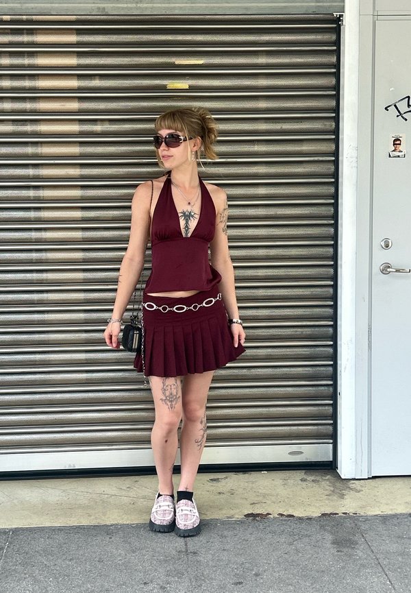 Top a halter bordeaux abbinato a una gonna plissettata, cintura in catena argentata e scarpe platform. Gli accessori includono occhiali da sole e una borsa piccola.