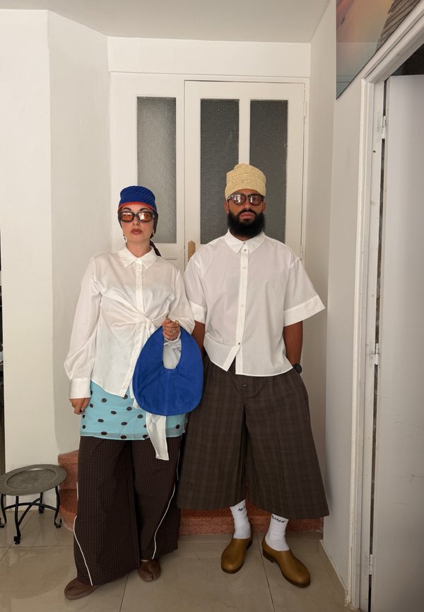 Deux personnes portant des chemises blanches, des pantalons larges, des bonnets en tricot, des lunettes de soleil et tenant un sac bleu se tiennent à l'intérieur devant une porte fermée.