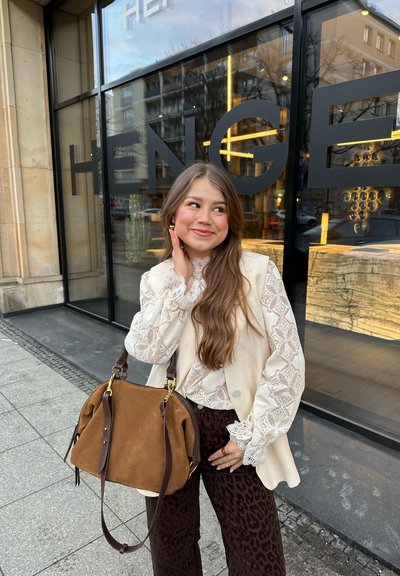 Joven mujer con cabello largo sonriente, vestida con una blusa de encaje, chaleco color crema, pantalones oscuros con estampado y sosteniendo un bolso grande de ante marrón, de pie frente a una tienda.
