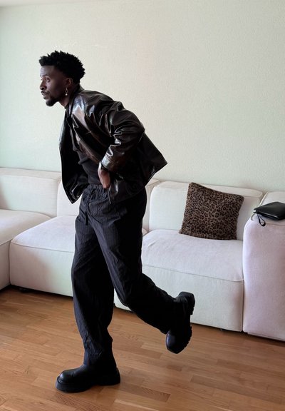 Hombre con chaqueta y pantalones de cuero negro, levantando una pierna en medio del paso, de pie sobre un suelo de madera junto a un sofá blanco con un cojín de estampado de leopardo.