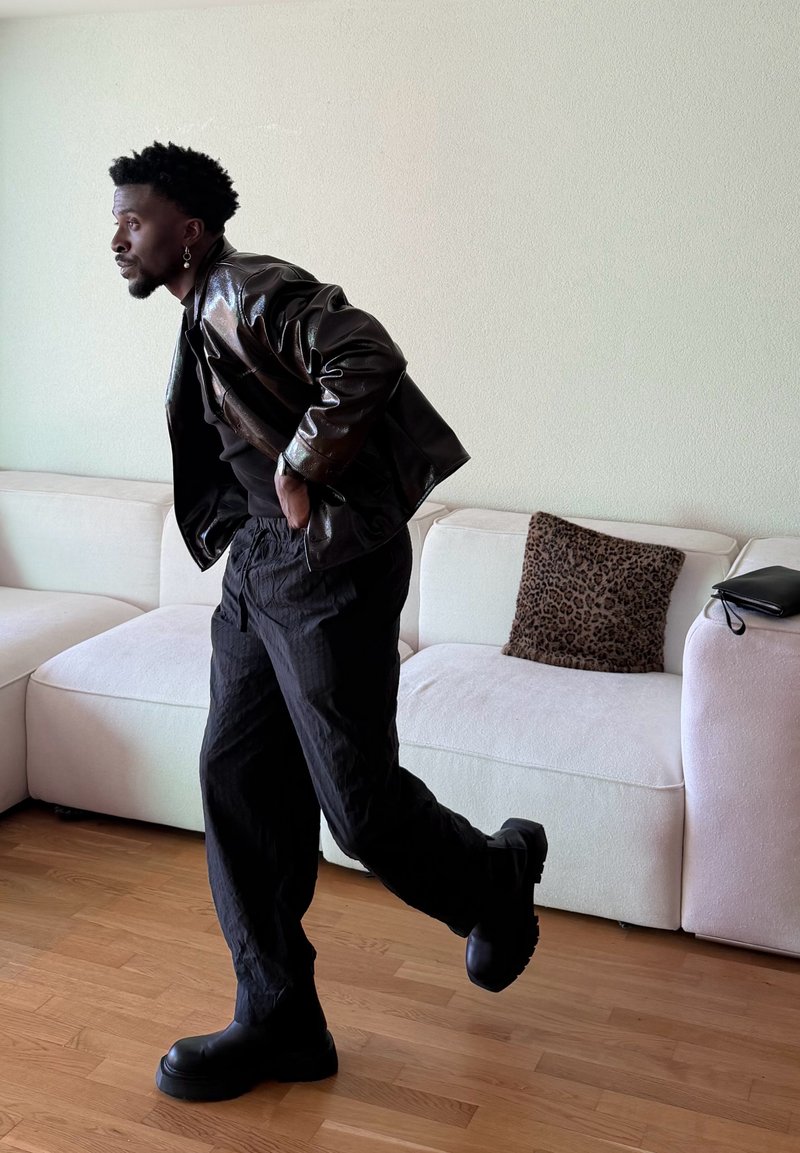 Homme portant une veste et un pantalon en cuir noir, levant une jambe en plein pas, debout sur un sol en bois à côté d'un canapé blanc avec un coussin imprimé léopard.