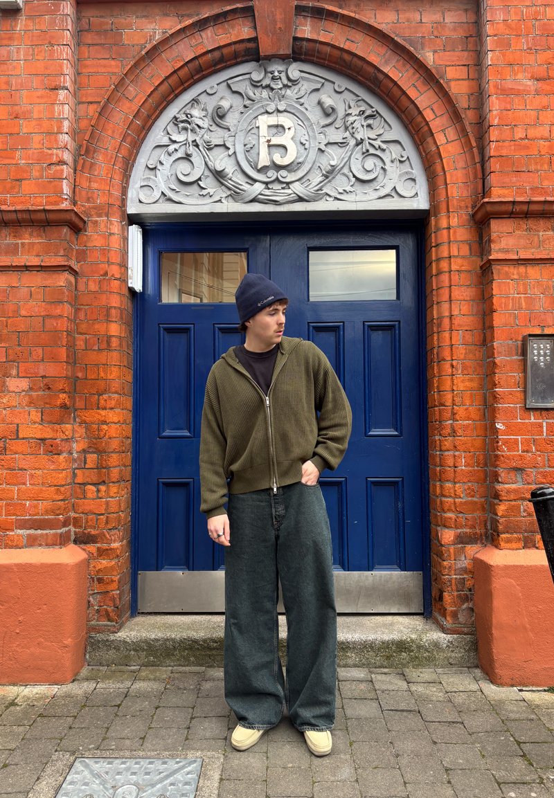 Jong persoon in een groene hoodie en brede jeans staat op een stenen straat voor een blauwe dubbele deur met decoratieve boog en de letter B erboven.