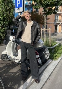 Jeune personne aux cheveux blonds portant une veste en cuir noire et un pantalon à jambes larges, debout et souriant près d'un scooter blanc sur le trottoir de la ville.